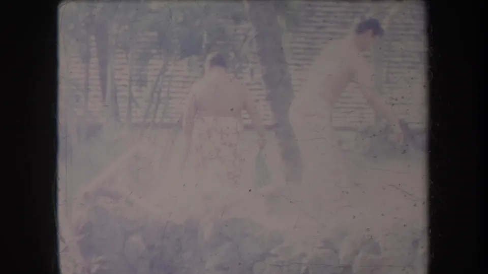 Two shirtless men appear to work near a stone wall in 1969 Hawaii, with steam or haze and a slatted structure behind them.