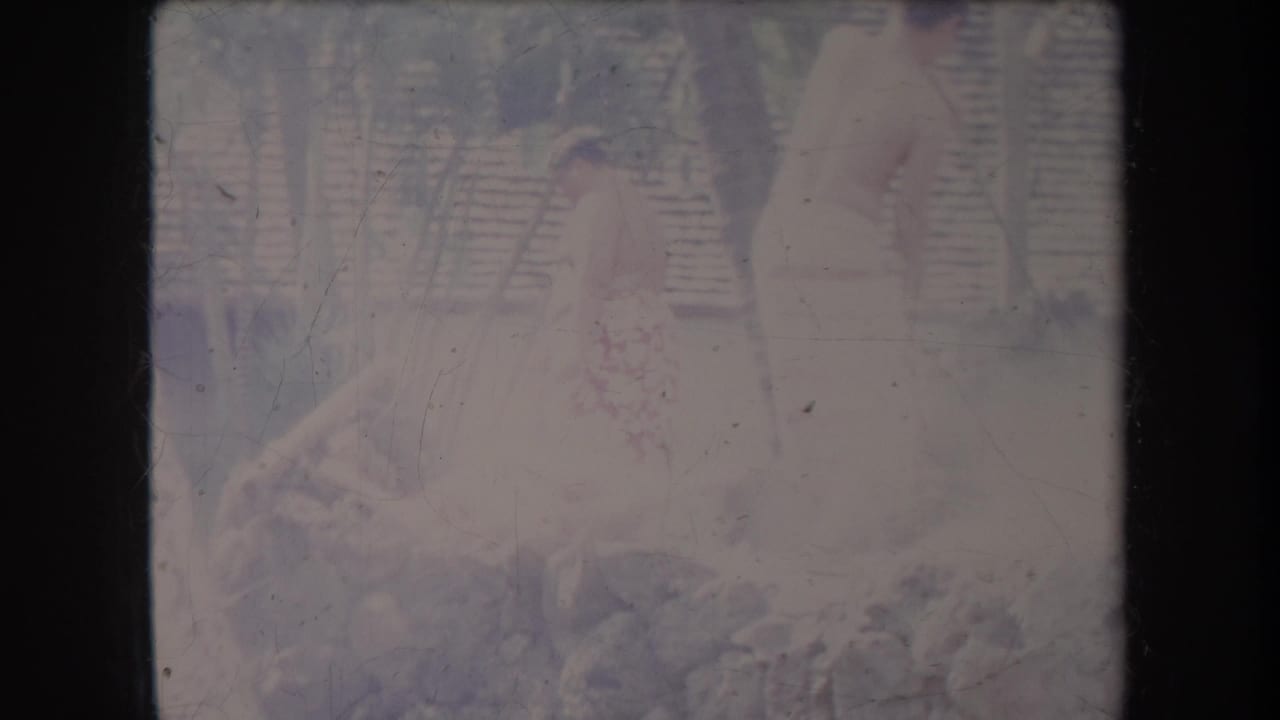 1969 Hawaii home-movie frame showing a person in light clothing near a rocky area with wood and a fence behind.