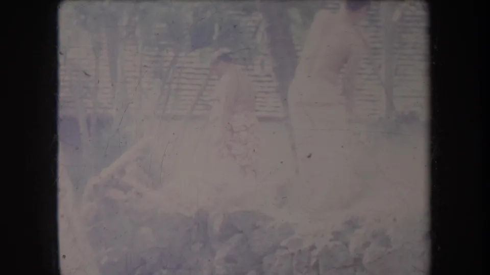 1969 Hawaii home-movie frame showing a person in light clothing near a rocky area with wood and a fence behind.