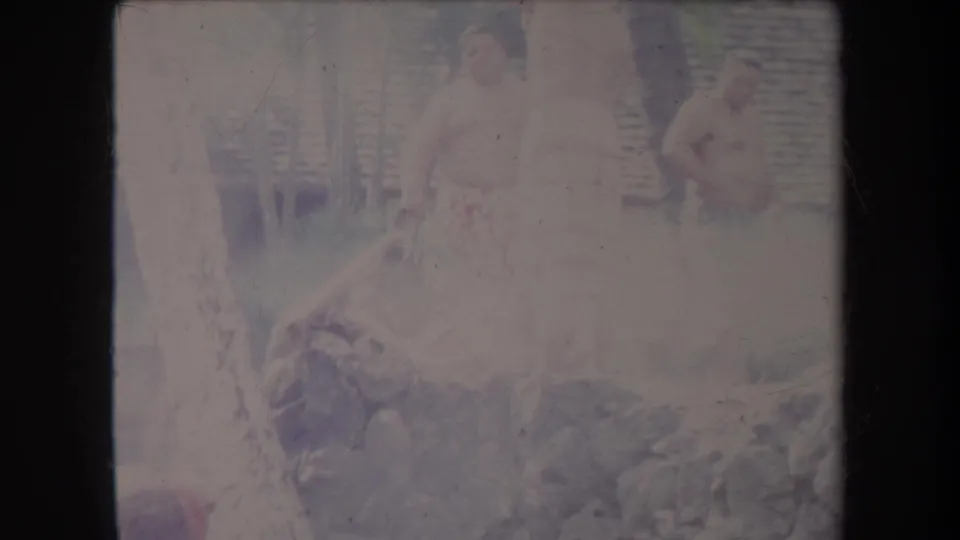 1969 Hawaii frame showing two adults in light clothing near rocks, wood, and a fence, with one wearing a patterned wrap.