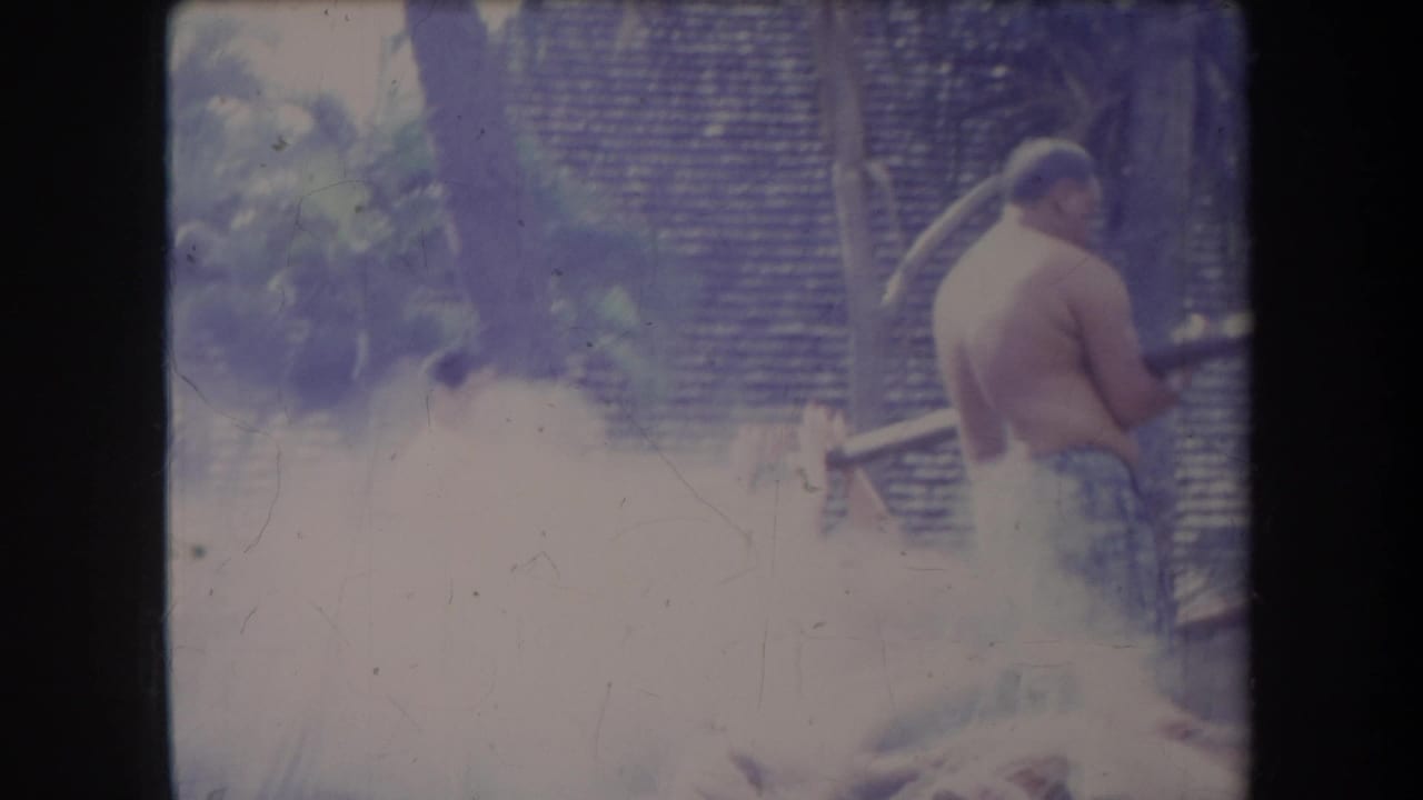 1969 Hawaii 8mm frame showing a shirtless man beside thick smoke rising from a pig roasting pit.