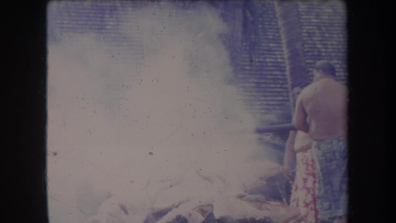 1969 Hawaii home-movie frame with dense smoke around an outdoor pig roast and a man standing at the right edge.