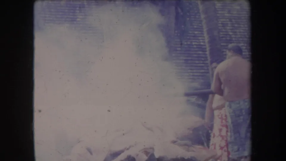 1969 Hawaii home-movie frame with dense smoke around an outdoor pig roast and a man standing at the right edge.