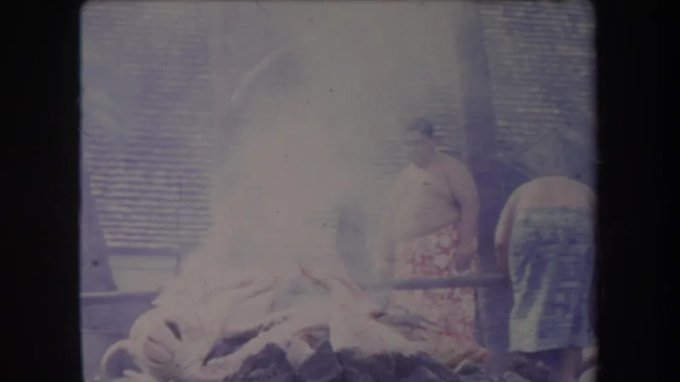 1969 Hawaii 8mm frame showing men at an outdoor pig roast, smoke rising around the pit and animal carcass.