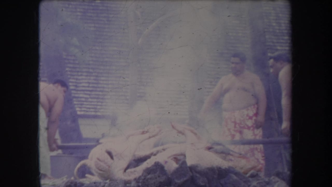 1969 Hawaii 8mm frame showing men near a whole pig roasting over an outdoor fire, with smoke and woven background panels.