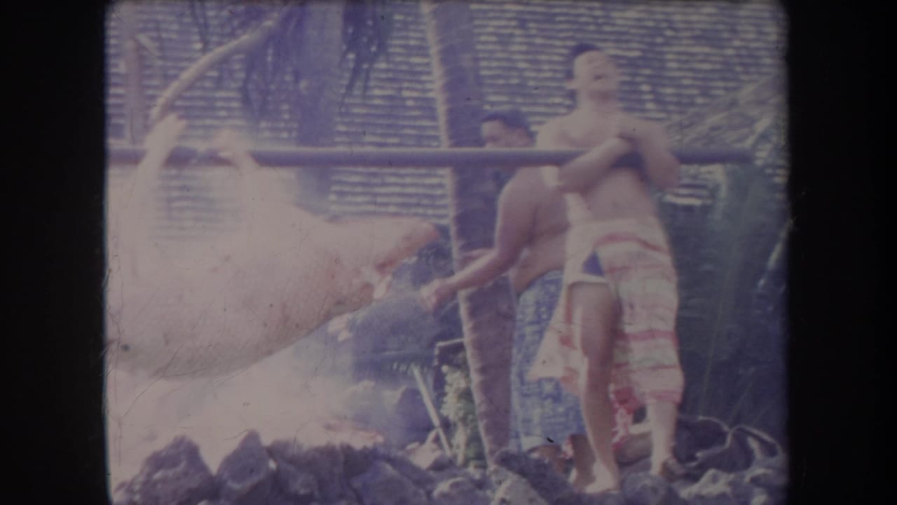 Men in patterned wraps stand beside a whole pig roasting over smoke and rocks.