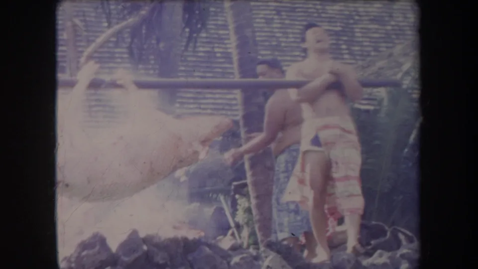 1969 Hawaii frame with men in wrap garments standing beside a whole pig on a spit over an open fire.
