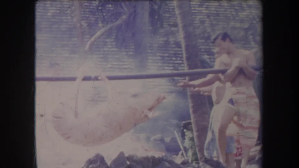 1969 Hawaii 8mm frame showing a man beside the pig roast, with smoke, rocks, palms, and a woven backdrop visible.