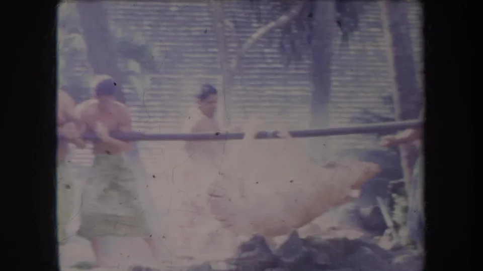 1969 Hawaii frame showing two men beside a smoking whole pig roast, with palms and woven panels in the background.