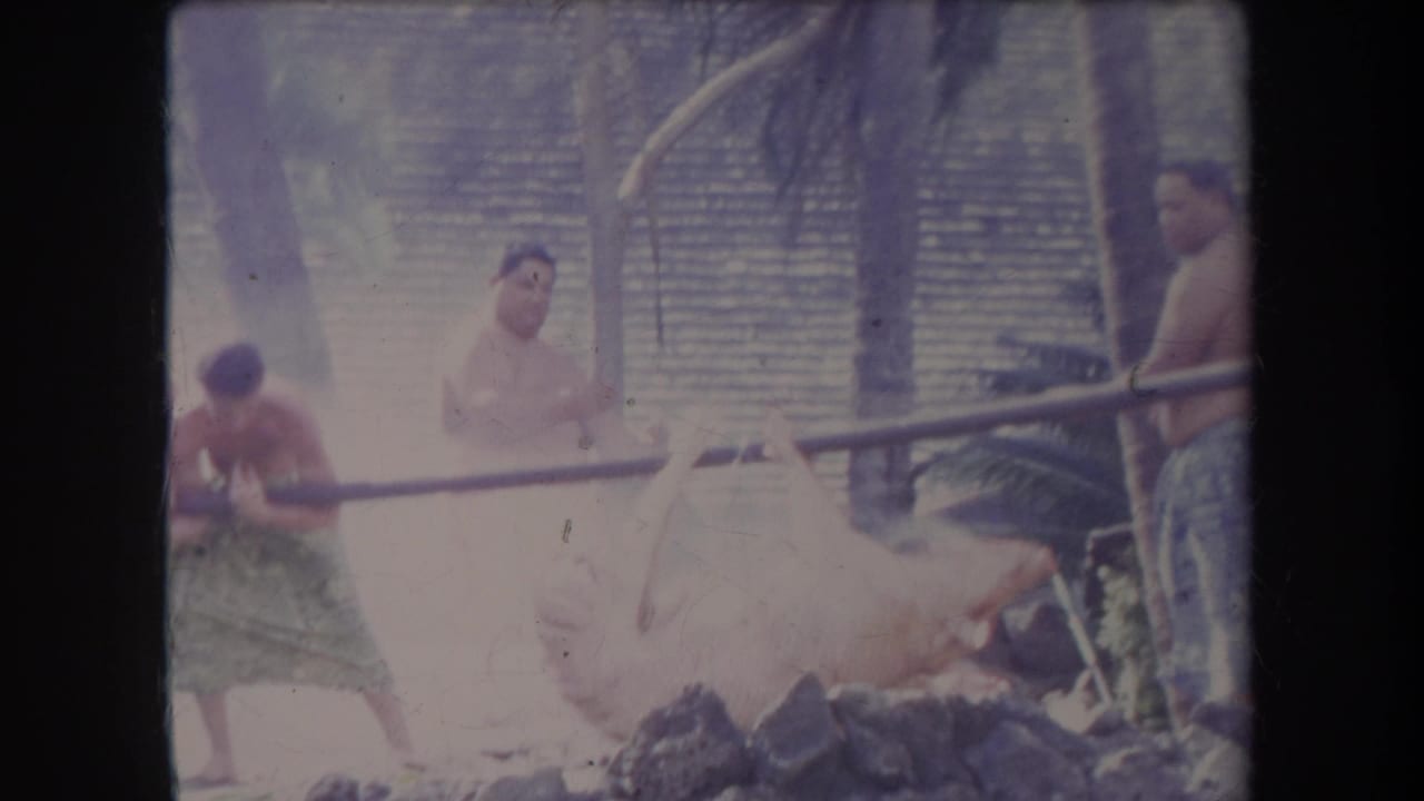 1969 Hawaii 8mm frame showing men in patterned wraps around a stone-lined pig roast pit with smoke, poles, palms, and a thatched wall.