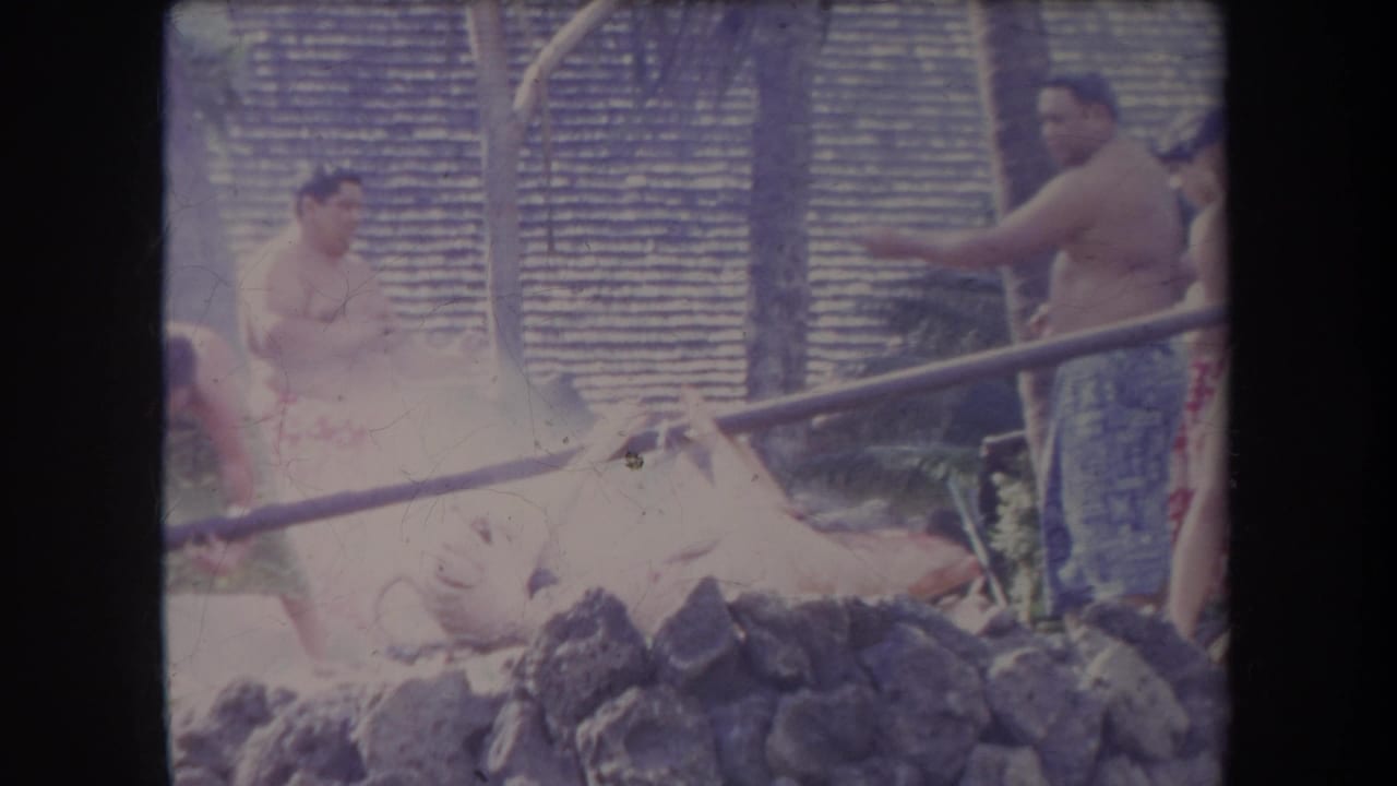 Several men stand near a whole pig over rocks, with poles and a thatched wall behind them.