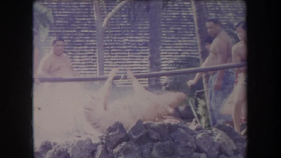1969 Hawaii 8mm frame showing a man in a light wrap by a smoking luau roast pit while others stand near the pig and stones.