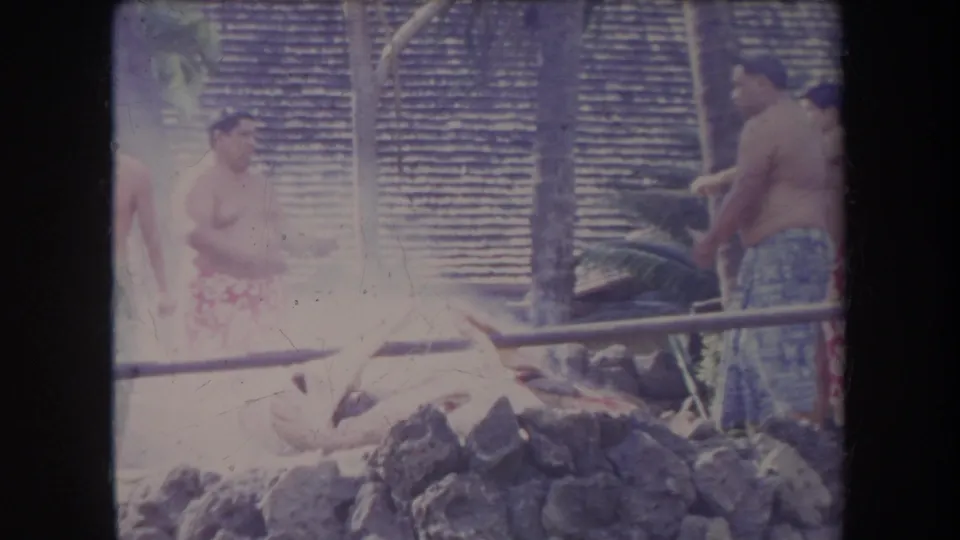1969 Hawaii home-movie frame of men beside a whole pig at a stone-lined luau pit, with poles, palm trunks, and a thatched wall visible.
