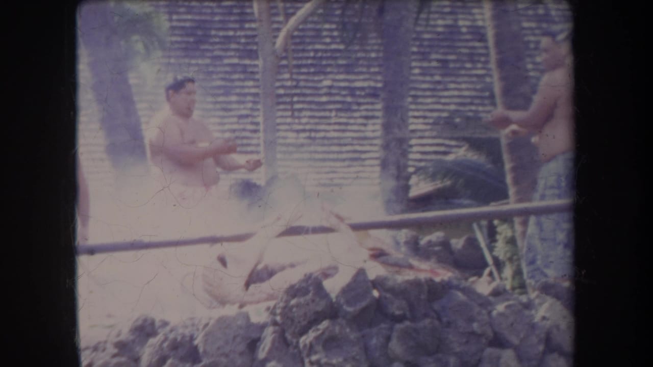 1969 Hawaii 8mm frame showing men outside near a smoking stone-lined cooking pit with tropical foliage and thatched material behind them.