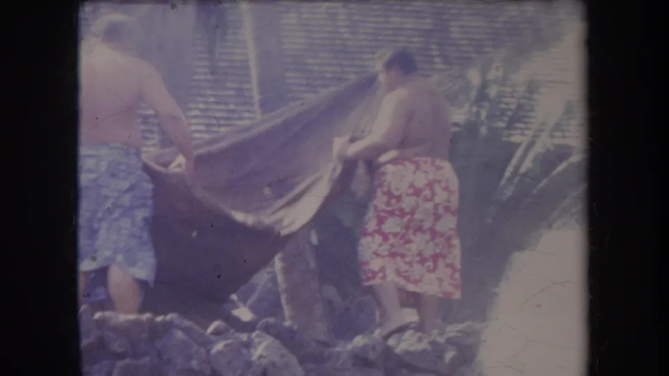 1969 Hawaii home-movie frame showing two adults handling a large blanket beside a stone-edged outdoor cooking area.