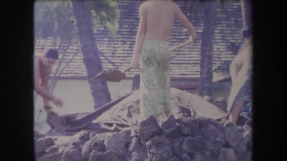 1969 Hawaii 8mm frame showing shirtless men in patterned wraps working around a covered, smoking pit near palms and a thatched background.