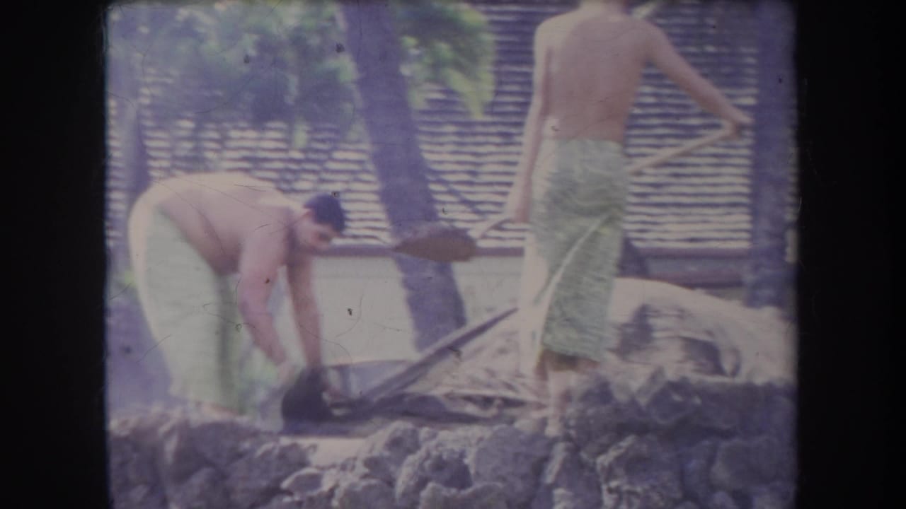 1969 Hawaii 8mm frame showing shirtless men in towels using shovels near rocks in a sunny tropical beach setting.