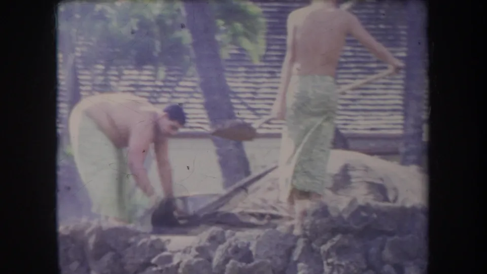 1969 Hawaii 8mm frame showing shirtless men in towels using shovels near rocks in a sunny tropical beach setting.