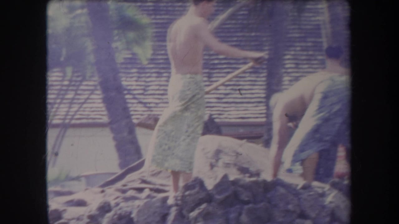1969 Hawaii home-movie frame with men in patterned towels working with shovels near rocks and a thatched-roof backdrop.