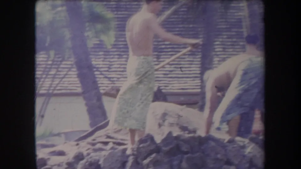 1969 Hawaii home-movie frame with men in patterned towels working with shovels near rocks and a thatched-roof backdrop.
