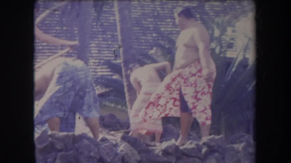 1969 Hawaii 8mm frame showing adults in towels clustered around rocks, with one person in a red patterned wrap visible.
