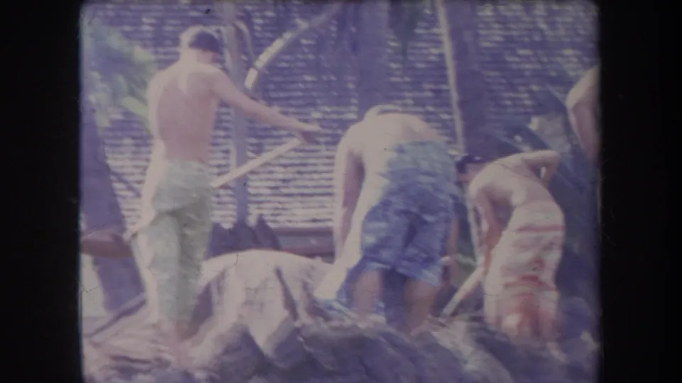 1969 Hawaii home-movie frame with shirtless men in towels digging or clearing rocks near palm trees and a thatched roof.