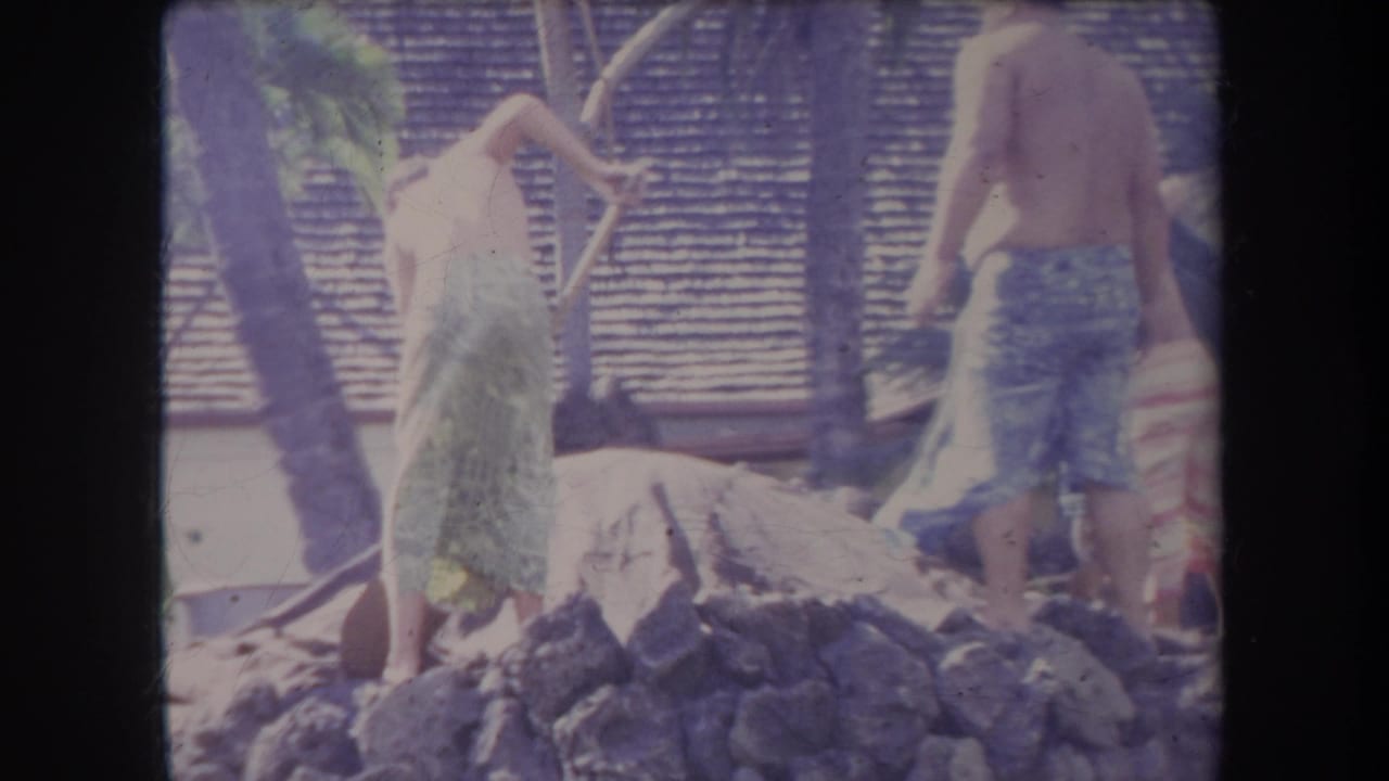 1969 Hawaii 8mm frame showing shirtless men near a rock pile and shovels in a tropical village-like setting.