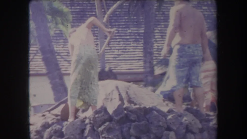 1969 Hawaii 8mm frame showing shirtless men near a rock pile and shovels in a tropical village-like setting.
