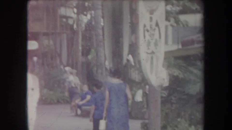 1969 Hawaii 8mm frame showing people in blue clothing walking past a carved sign in a tropical pedestrian area.