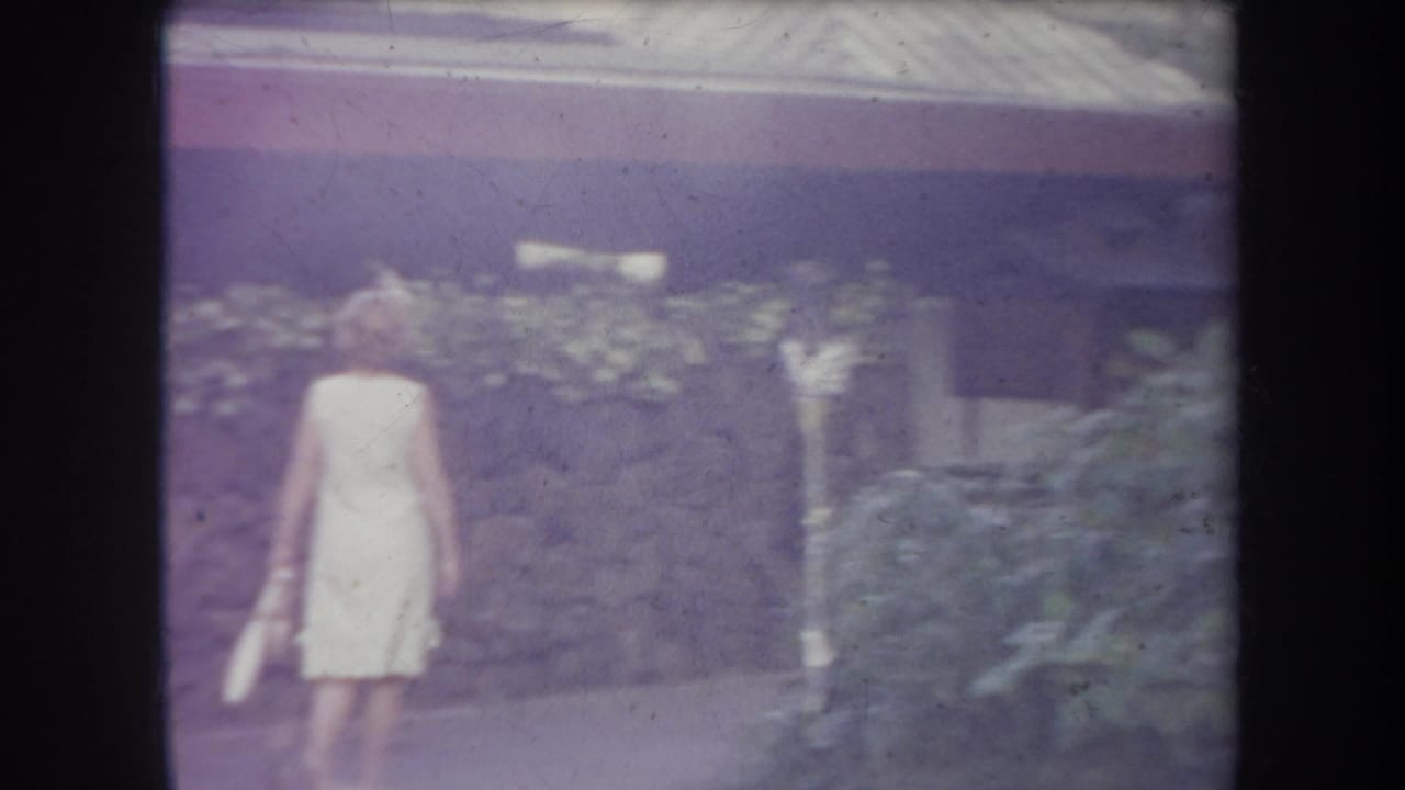 Woman in a pale dress walking past a stone wall and foliage.