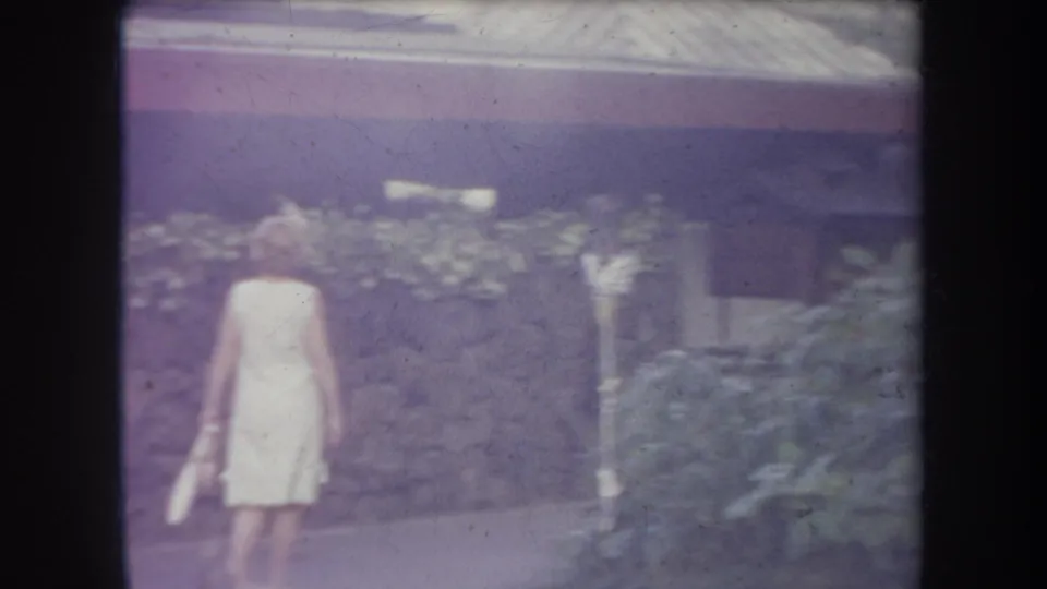 Woman in a light dress walks through a landscaped Hawaii theme park area in 1969 home-movie footage.