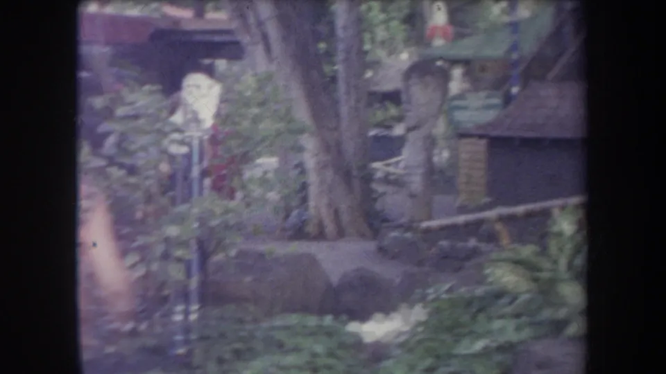 A landscaped theme park garden with a decorative lamp post, dense plants, and a small structure in 1969 Hawaii footage.