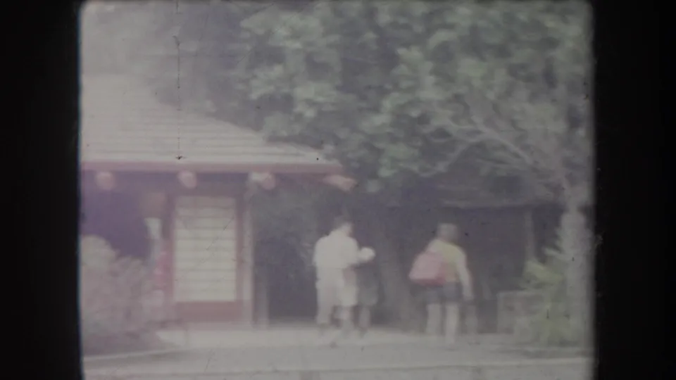 1969 Hawaii frame showing two adult visitors near a small garden structure beneath dense tropical foliage.