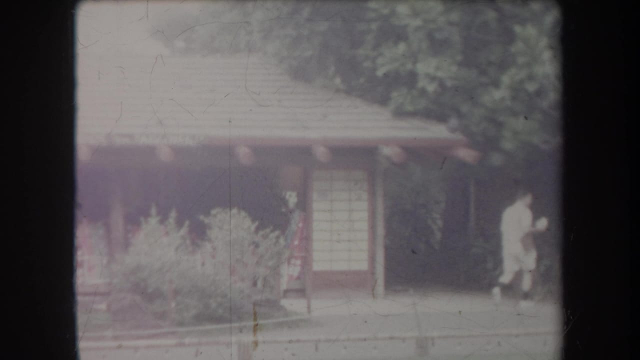 1969 Hawaii home-movie frame showing visitors near a low garden building, sliding-style doors, and landscaped plants.