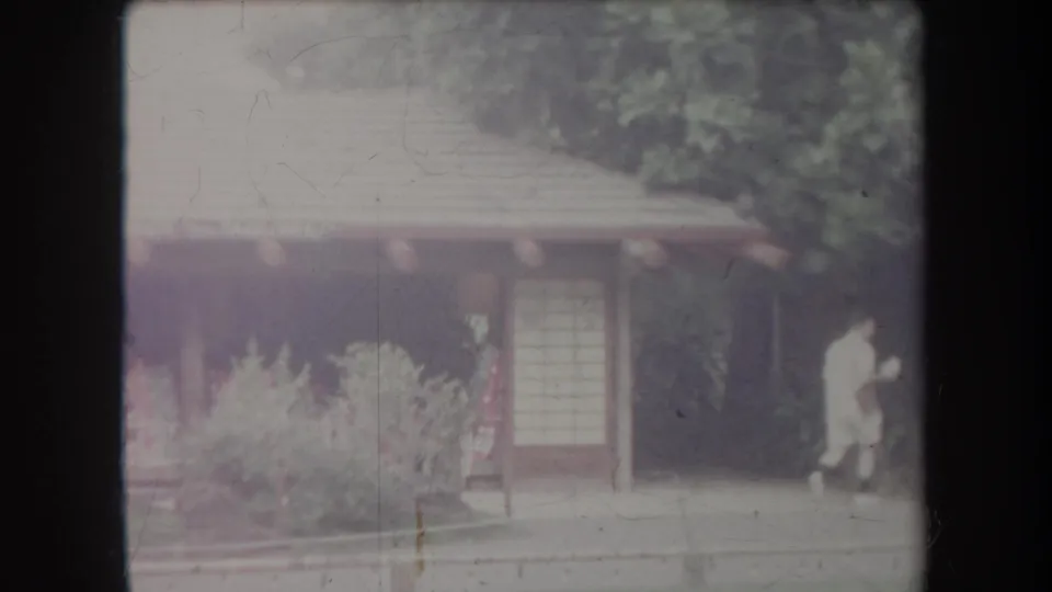 1969 Hawaii home-movie frame showing visitors near a low garden building, sliding-style doors, and landscaped plants.