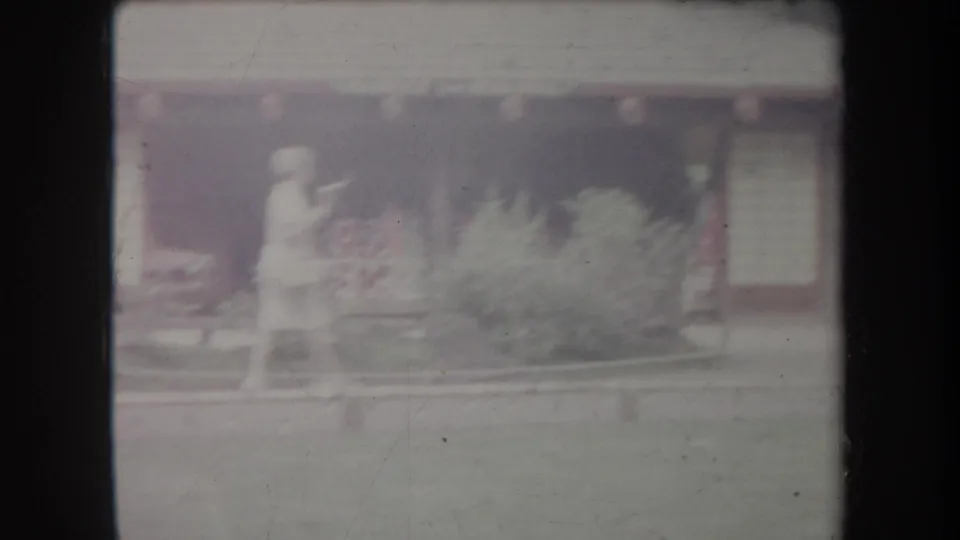 Soft 8mm frame from Hawaii showing a person by a planted garden bed in front of a shaded park building.
