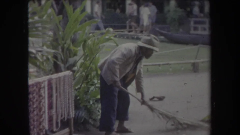 1969 Hawaii park scene with a hat-wearing groundskeeper sweeping a walkway near tropical foliage and a lawn.