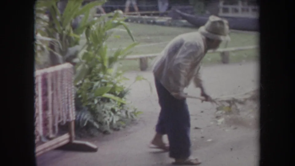 Faded 8mm Hawaii frame showing a groundskeeper sweeping beside tropical plants, a table, and a green lawn.