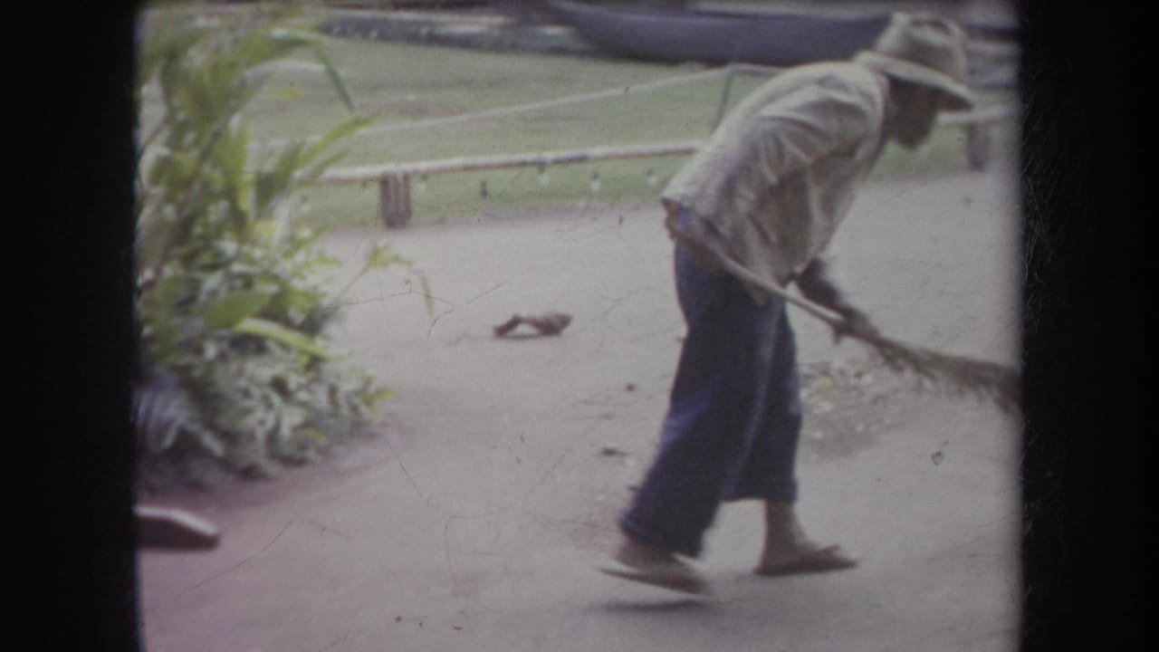 In 1969 Hawaii home-movie footage, a man in work clothes sweeps pavement beside plants and a low wooden railing.