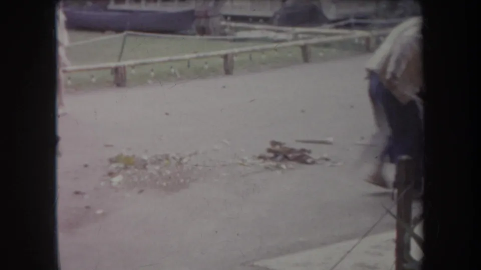 A handheld 8mm frame in Hawaii shows the man partly cropped as swept leaves and debris gather on the paved walkway.