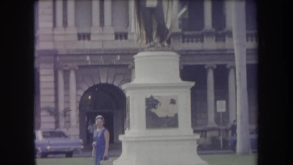 In 1969 Hawaii, a woman in a blue dress stands by a large white monument pedestal before a classical-style building and parked car.