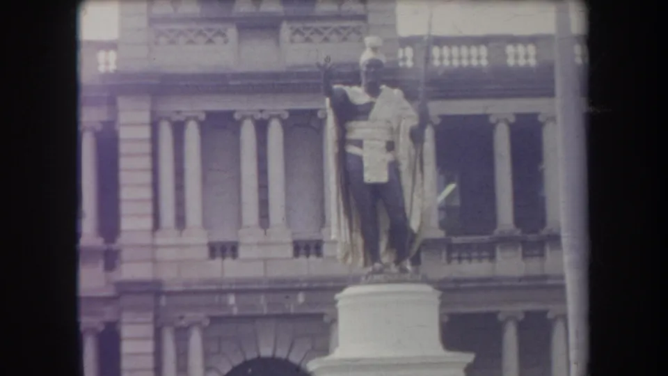 A standing statue in traditional-looking attire rises from a white pedestal before a columned public building in 1969 Hawaii footage.