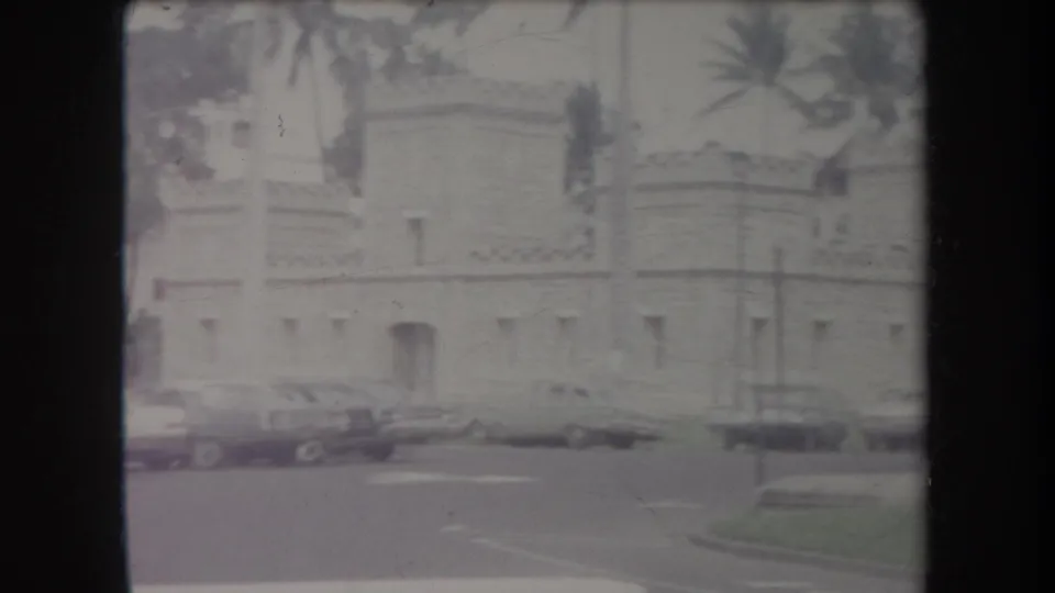 1969 Hawaii home-movie frame showing a stone building across a street with parked cars, palms, and soft faded color.