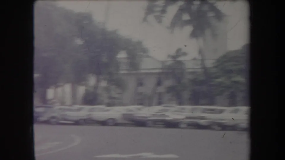 1969 Hawaii frame showing period cars parked along a curved street, with trees and a large building softened by motion and grain.
