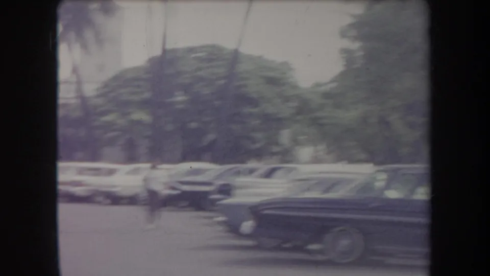 1969 Hawaii 8mm frame showing parked mid-century cars beside a tree-lined street, with palm trunks and soft motion blur.