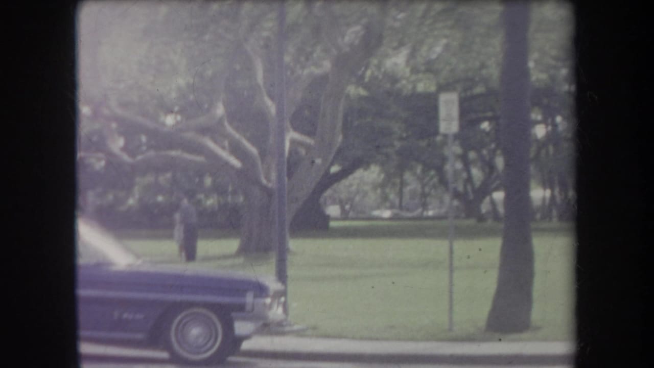 1969 Hawaii home-movie frame with a purple classic car at the curb, open grass, mature trees, and a small distant person.