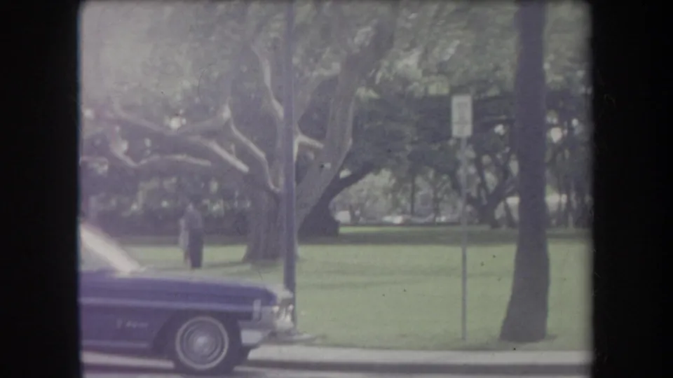 1969 Hawaii home-movie frame with a purple classic car at the curb, open grass, mature trees, and a small distant person.