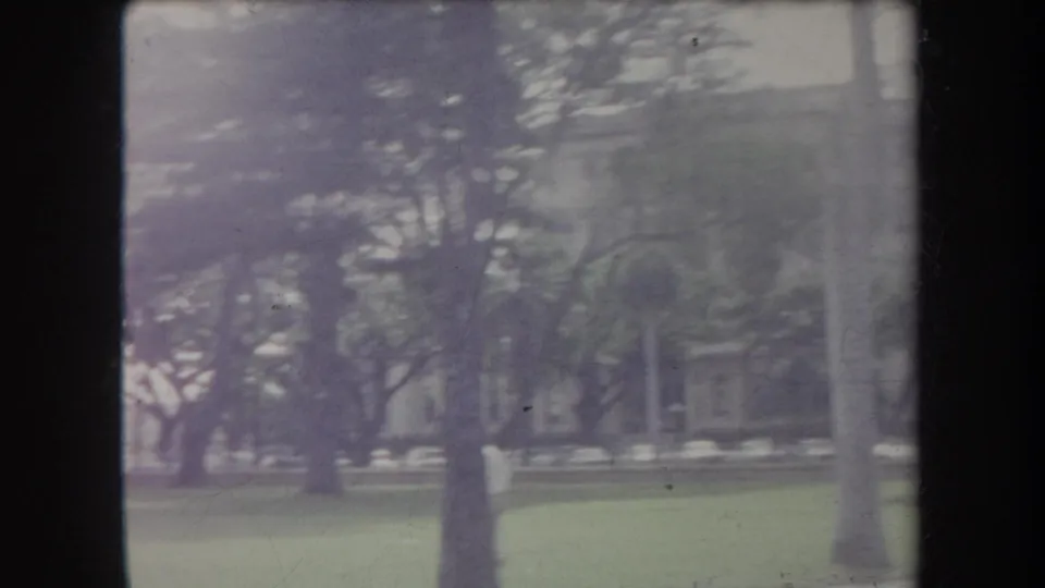 1969 Hawaii home-movie frame of trees and lawn with a partially obscured stone or concrete building and arched windows beyond.