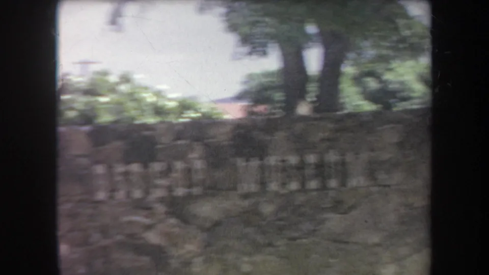 1969 Hawaii frame with a weathered stone wall or sign, faint lettering, tropical planting, trees, and bright overexposed sky.