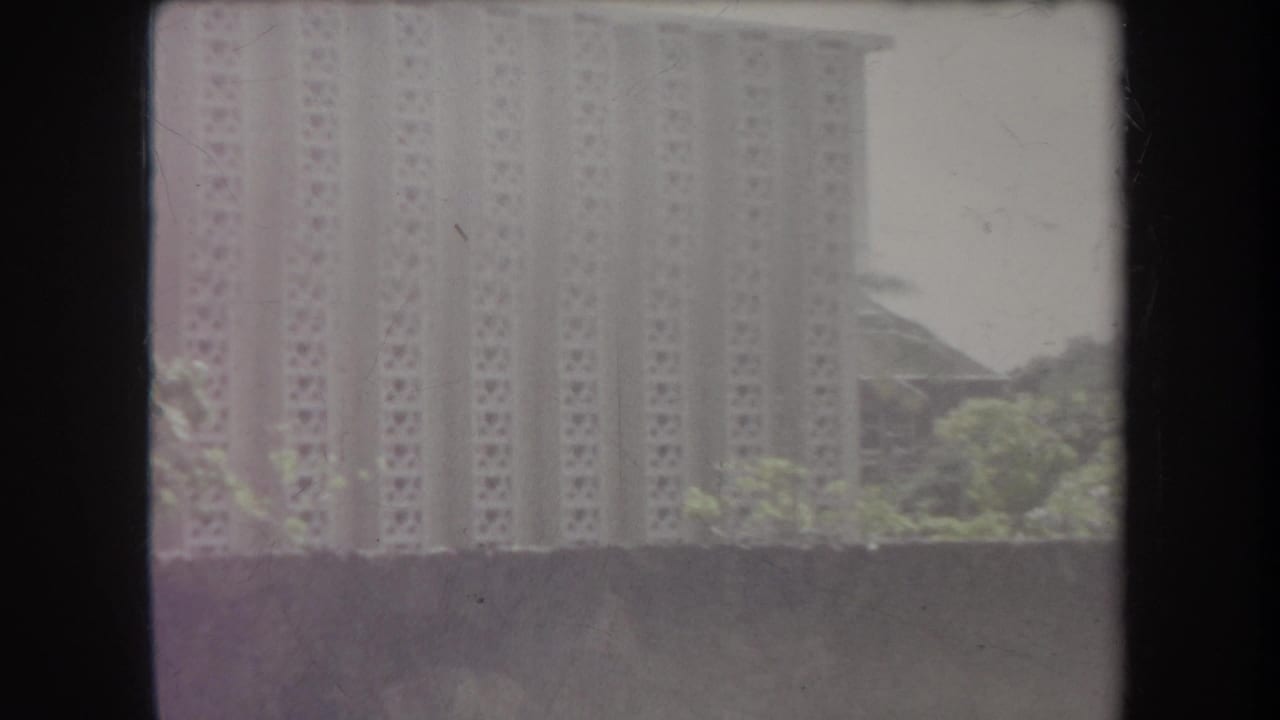 1969 Hawaii home-movie frame showing a tall building beyond trees and a wall, with soft focus and dark 8mm frame borders.